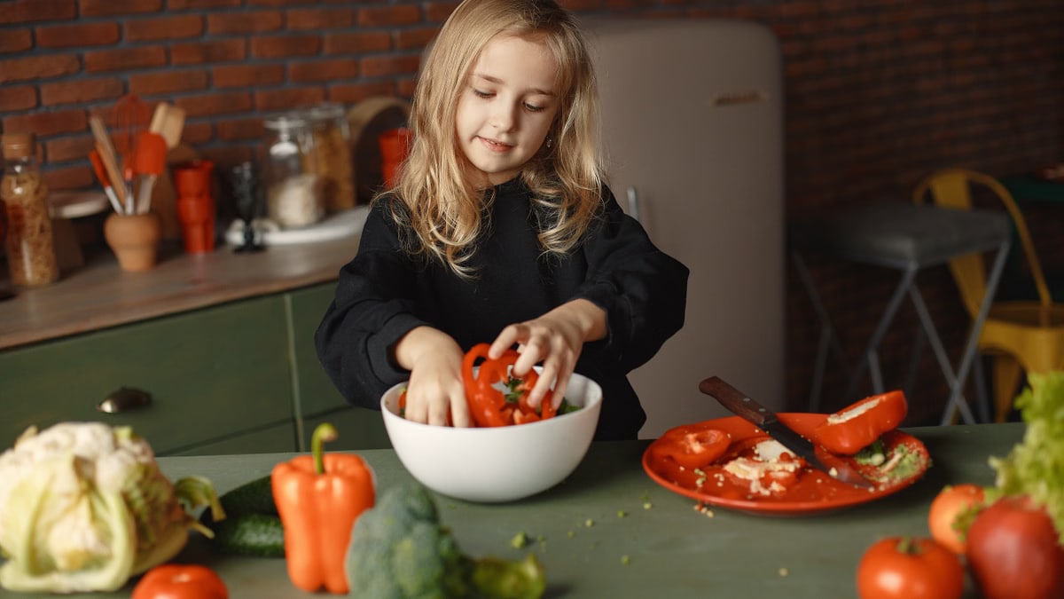 Content little kid in casual clothes preparing vegetable salad and putting ingredients into deep ceramic bowl while standing in loft style kitchen at home